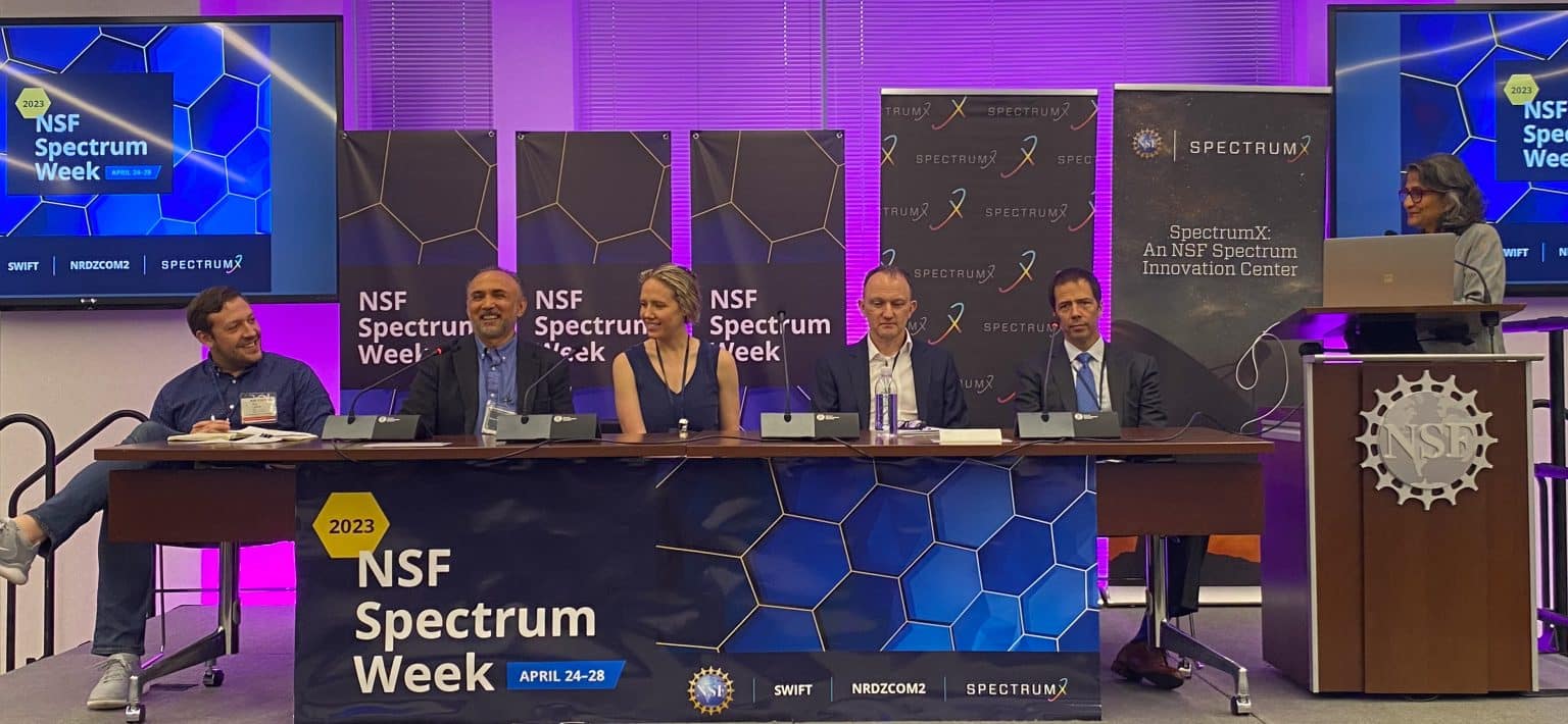 This image is from a panel during the SpectrumX center meeting at 2023 NSF Spectrum Week. There are five panelists at a table with microphones, with a host at the podium